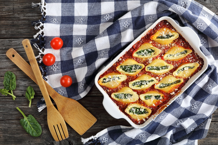 shell shape pasta stuffed with creamy soft cheese and spinach sprinkled with parmesan cheese in baking dish with kitchen towel and wooden spatulas on table, healthy dinner recipe, view from aboveの写真素材