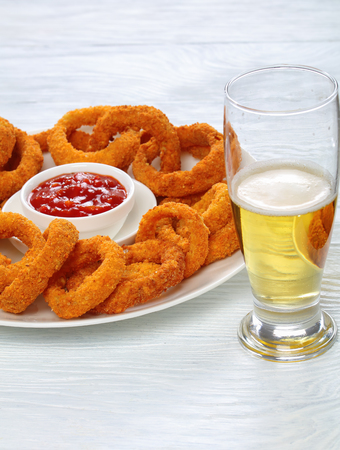 delicious golden battered, breaded and deep fried crispy onion rings served on white platter with tomato sauce in center and glass of beer on table, side view from above, close-upの写真素材