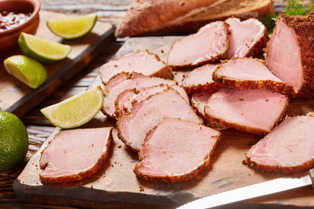 close-up of delicious sliced ham seasoned with ground spices served on wooden cutting board with barbecue sauce, lime slices and rye bread at background, view from aboveの写真素材