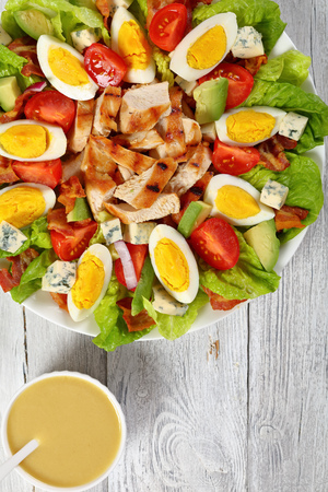 american cobb salad of hard boiled egg, blue mold cheese, crispy fried bacon, romaine lettuce, grilled chicken breast, tomato and avocado chunks on a plate. dressing in bowl on tableの写真素材