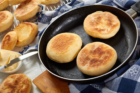 cooking breakfast - baking english muffins on a skillet and are chilling on a grid on wooden table with kitchen towel, fresh butter and knife, view from above, close-upの写真素材