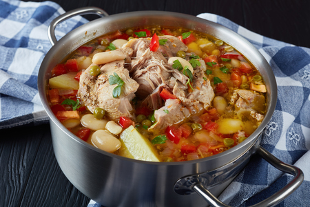 delicious rich pork vegetable soup with white beans, green peas, red bell pepper, herbs, species in a metal casserole on a black wooden table, view from above, close-upの写真素材