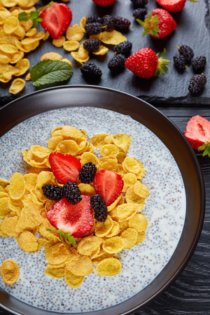 Corn flakes with chia seeds pudding, fresh strawberry,and mulberries in a bowl on black wooden table with ingredients on cutting board, vertical view from above, close-up,の写真素材