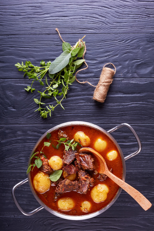 delicious Beef Stew with tender cubes of meat, whole new potatoes, carrots and herbs in a metal pot on black wooden table with bouquet of aromatic herbs, vertical view from aboveの写真素材