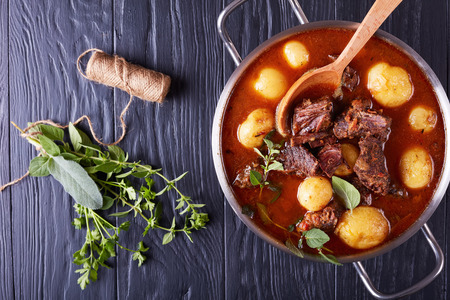 Beef Stew with tender cubes of meat, whole new potatoes, carrots and herbs in a metal pot on black wooden table with bouquet of aromatic herbs, view from above, close-upの写真素材