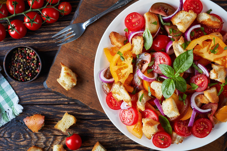 delicious fresh classic italian salad panzanella with tomatoes, croutons basil and onion rings on a white plate on an old wooden table with ingredients, view from above, flat lay, close-upの写真素材