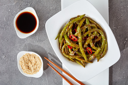 Spicy Chinese stir-fried Green Beans in a white bowl with chopsticks on a white rectangular plate on a concrete table with soy sauce and sesame seeds, view from above, close-up, flat lay,の写真素材