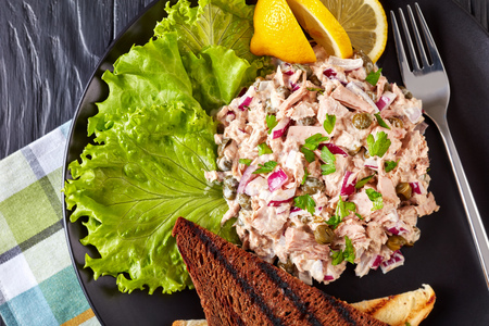 close-up of Tuna, capers and red onion salad served with toasted bread, lemon wedges and lettuce leaves on a black plate on a wooden table with napkin, view from above, flat layの写真素材