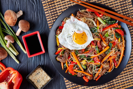 Sweet and savory  korean Japchae: bouncy cellophane noodles with mixed crunchy vegetables: spinach, red bell pepper, onion, carrot, garlic, shiitake mushrooms and beef with fried egg on top, flat layの写真素材