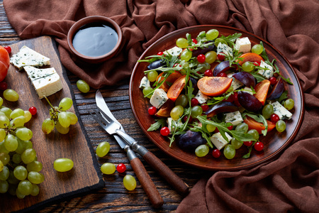 delicious autumn salad with chocolate persimmon, blue cheese, green grapes, plum slices and arugula on a earthenware plate with ingredients on a cutting board, view from above, close-upの写真素材