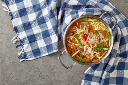 overhead view of duck noodle vegetables soup in a metal casserole on a concrete table with kitchen...