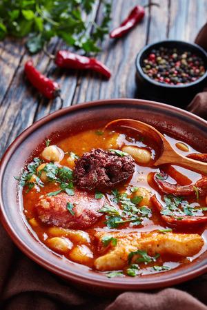 Fabada Asturiana Bean hearty Stew in an earthenware bowl with spoon on a rustic wooden table with brown cloth, chili peppers and parslaey, spanish cuisine, vertical view from above, close-upの写真素材