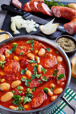 hearty beans stew with sausages, herbs and spices in tomato sauce in a metal casserole on a concrete table with ingredients, fasolka po bretonsku, comfort food, vertical view from aboveの写真素材