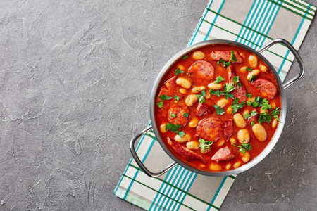 hearty beans stew with sausages, herbs and spices in tomato sauce in a metal casserole on a concrete table with napkin, fasolka po bretonsku, polish cuisine, view from above, flatllay, copy spaceの写真素材