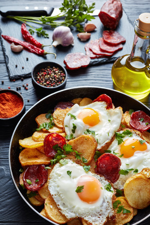 delicious fried sunny side up eggs with potatoes, thinly sliced pork sausages in a skillet on a black tabletop with ingredients at the background, european cuisine, vertical view from aboveの写真素材