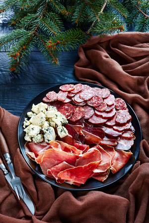 overhead view of a christmas antipasti - cheese, sausages and dry cured meat - jamon,  chorizo, blue mold cheese, sliced fuet sausages on a plate on a table with fir tree, view from above, flatlayの写真素材