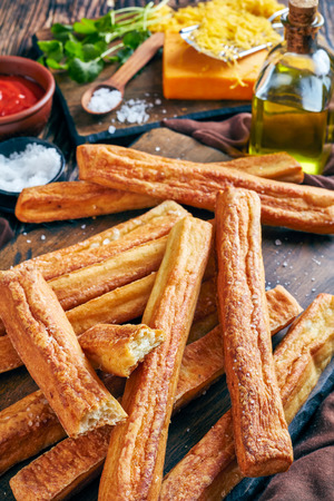 overhead view of Cheese Straws, Cheesy Bread Sticks on a cutting wooden board with grated cheddar cheese, tomato sauce, olive oil on a table, vertical view from above, close-upの写真素材