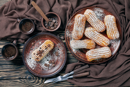 overhead view of delicious Homemade Eclairs sprinkled with powdered sugar and chocolate drops on an earthenware plate on a dark rustic table with cups of coffee, view from above,  flatlayの写真素材