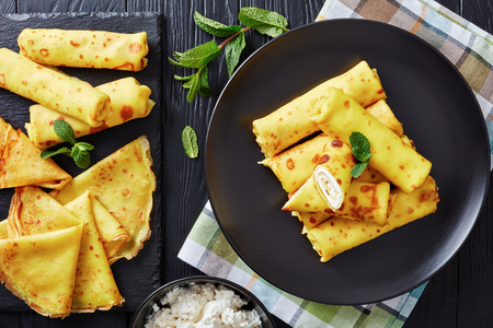 Crepe Roll Ups with mascarpone filling on a black plate on an old wooden table with fresh mint leaves and ingredients at the background, view from above, flatlay, close-upの写真素材