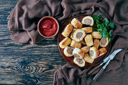 sausage rolls on an earthenware plate on a dark wooden rustic table with brown cloth and tomato...