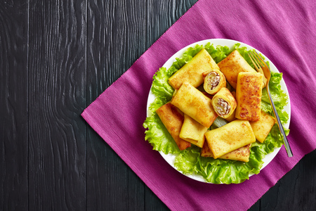 overhead view of Savory crepe rolls with ground chicken meat and champignon filling served on a bad of fresh lettuce leaves on a white plate on a black wooden table, flatlayの写真素材