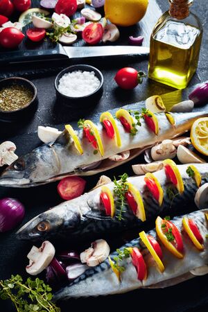 three whole raw mackerel with lemon, tomatoes, mushrooms, spices and herbs. scomber on a stone tray on a concrete table, vertical view from above, close-upの写真素材