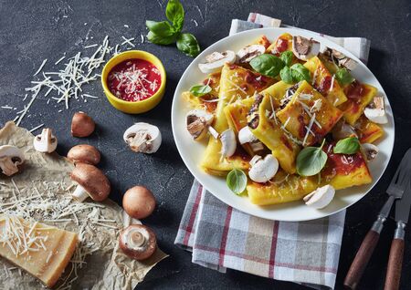 rolls of crepes with parmesan and mushrooms filling on a white plate on a concrete table with tomato sauce, piece of cheese and kitchen towelの写真素材