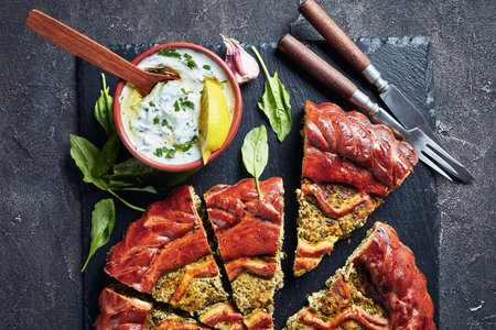 close-up of sliced Spinach Ricotta Pie on a black slate tray with tzatziki sauce in a bowl on a...