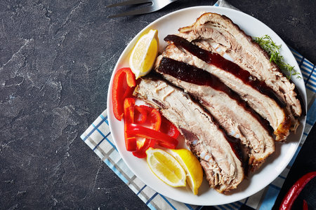 sliced roast pork belly on a white plate on a concrete table, horizontal view from above, flatlay, empty space, close-up