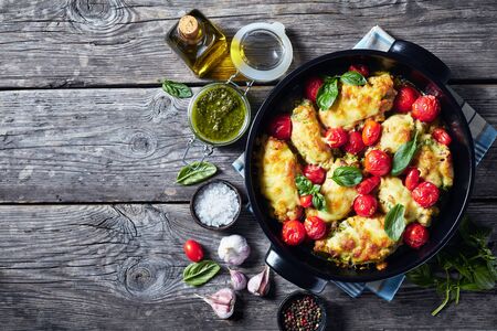 baked Chicken breast rolls filled with basil pesto, tomatoes and cheese in a black ceramic baking dish on a rustic wooden tableの写真素材