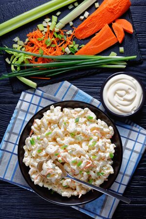 macaroni salad of elbow pasta with shredded carrots, celery, spring onions and mayonnaise dressing in a black bowl on wooden table. ingredients on a black stone board, vertical viewの写真素材