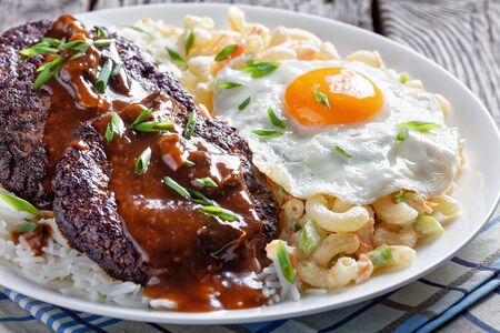loco moco of white rice, topped with a hamburger patty, a fried egg, brown gravy, and macaroni salad served on a white plate on a rustic wooden table, Hawaiian cuisine, view from above, の写真素材
