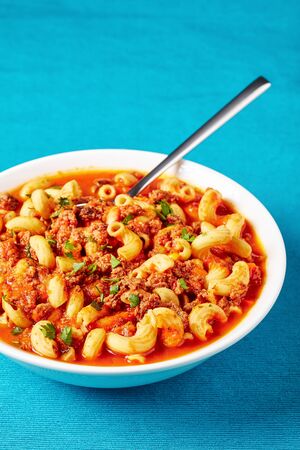 portion of goulash of elbow pasta, beef, celery and tomatoes served in a white bowl on a blue textile mat, view from above, close-upの写真素材
