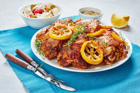 crispy fried juicy Chicken Thighs With Roasted Lemon Slices On a white plate on a concrete table with Mediterranean salad in a bowl, view from aboveの写真素材