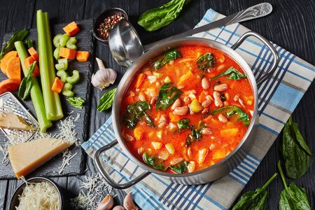 borlotti beans soup with celery, vegetables, spinach, parmesan and tomatoes in a metal saucepan on a black wooden table, ingredients on a black stone board, horizontal view from aboveの写真素材