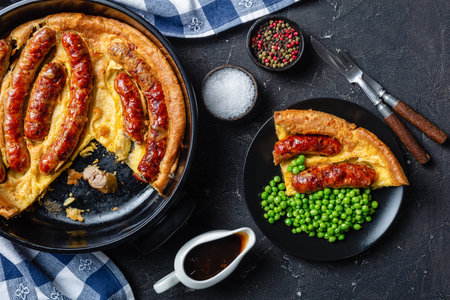 close-up of Toad in the hole, Sausage Toad, sausages in Yorkshire pudding batter in black ceramic baking dish and a portion on a plate with green peas and onion gravy,  view from above, flatlayの写真素材