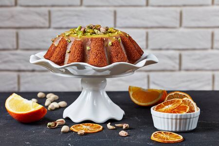 pumpkin carrot sponge cake with orange glaze, sprinkled with pistachios on a white ceramic stand with a brick wall at the background and ingredients on a table, close-upの写真素材