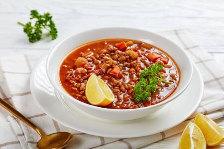 close-up of lentil soup with vegetables and lemon wedges in a white bowl on a wooden table with a golden spoon and toasts, horizontal view from aboveの写真素材