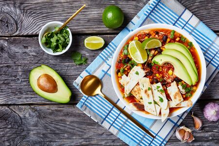 Chicken Taco Soup with black bean, corn kernels, topped with tortilla strips, avocado slices and lime in white bowl on a rustic wooden table, horizontal view from above, flat lay, copy spaceの写真素材