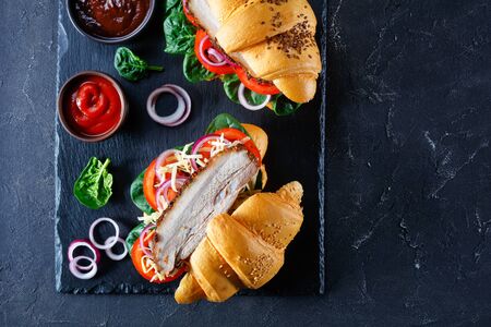 Croissant sandwiches with pork, tomatoes, fresh spinach, onion and shredded cheese on black stone board over stone textured grey background, flat lay, close-up, horizontal view from aboveの写真素材