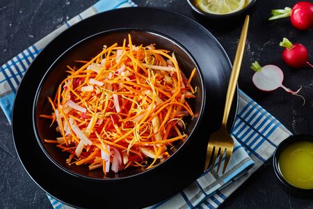 close-up of Fresh carrot radish and apple salad sprinkled with quinoa seeds and lemon zest in a black bowl on a wooden table with homemade dressingの写真素材