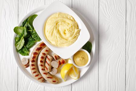 Cheesy whipped potato aligot with grilled pork sausages dijon mustard and fresh baby spinach, lemon wedges served on a white serving tray on a white wooden table, top view, close-upの写真素材