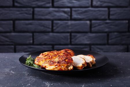 Baked chicken breasts with spices served on a black plate with golden cutlery and rosemary sprigs, on a dark concrete background in front of black brick wall, horizontal orientation, copy spaceの写真素材