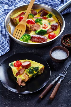 frittata with sausages, broccoli, cherry tomatoes, melted cheese in a skillet on a concrete table and a portion on a plate, vertical view from above, close-upの写真素材