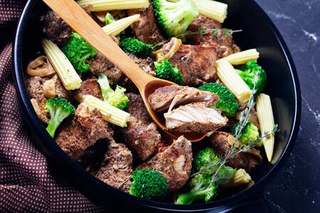 Leftover steak kebabs with vegetables on a black baking dish: broccoli, corn, onion, and garlic on a dark concrete background, close-up, horizontal orientationの写真素材