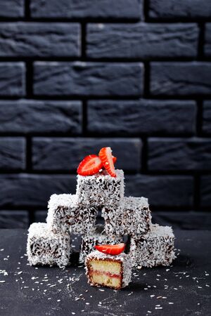 Vanilla sponge mini cakes with strawberry jam layer inside, coated with chocolate and coconut on top on a dark concrete tableの写真素材