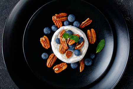 Uncooked brie cheese with blueberries, pecans with mint on top on a black plate on a dark concrete