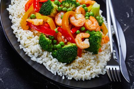 close-up of a portion of fried shrimps with bell pepper, onion, broccoli and green peas on steamed rice on a black plate on a concrete tableの写真素材