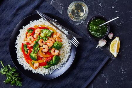fried shrimps with bell pepper, onion, broccoli and green peas on steamed rice on a black plate with a glass of white wine on a concrete table, view from above, flat layの写真素材