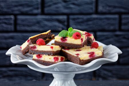 delicious vegetarian chocolate cheesecake bars with raspberries on a white cake stand with a brick wall at the background, horizontal view, close-upの写真素材